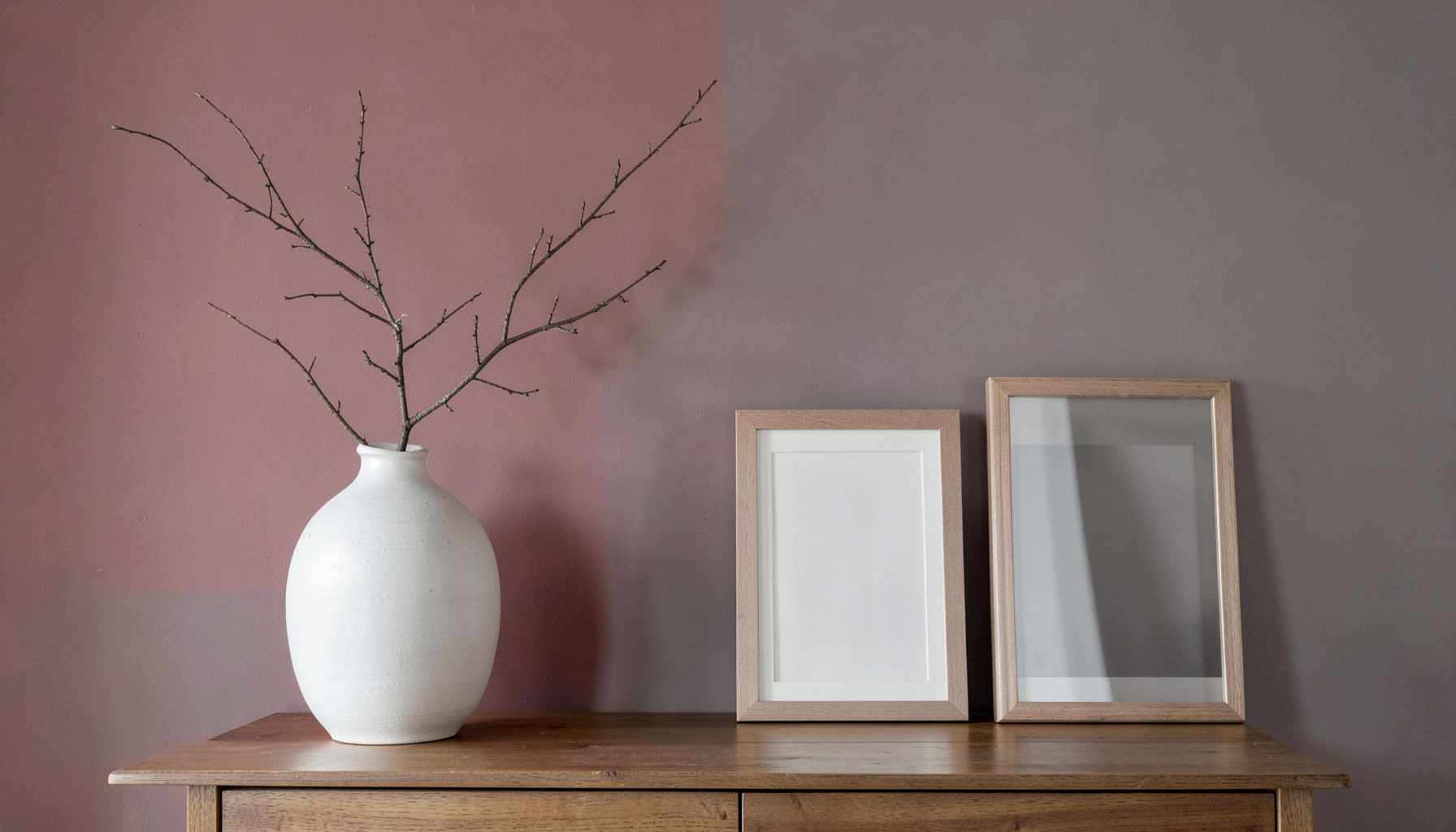 Interior wall in muted red and gray tones with decorative white vases, a slender branch, and empty picture frames on a sideboard, evoking calm and thoughtful simplicity